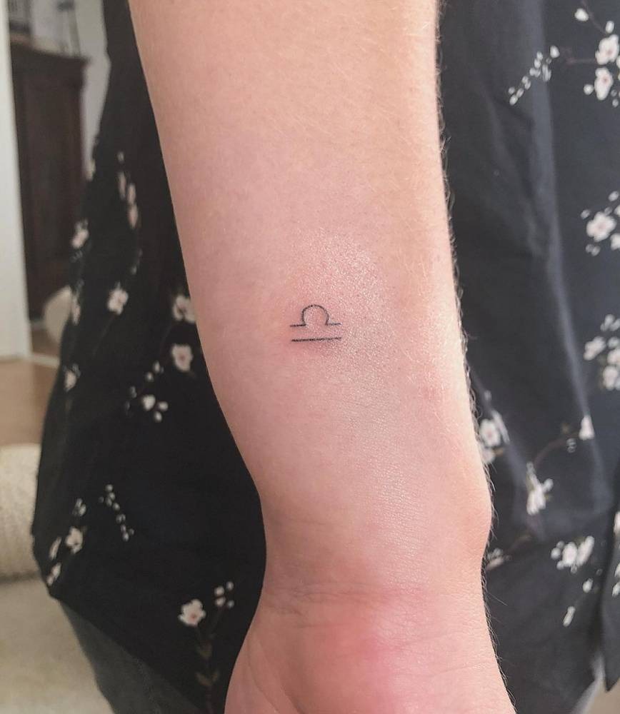 This tattoo features a delicate and minimalist depiction of the Libra symbol, placed on the wrist.