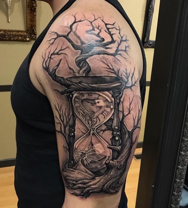 This shoulder tattoo showcases an hourglass intertwined with the roots and branches of a tree, representing the adventure of life and its cyclical nature.
